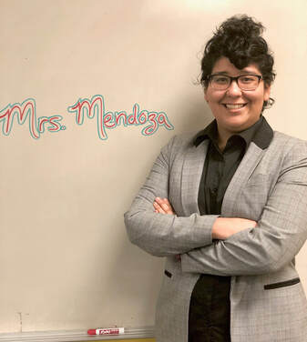 Mrs. Mendoza in a gray jacket standing in front of her whiteboard where her name is written.